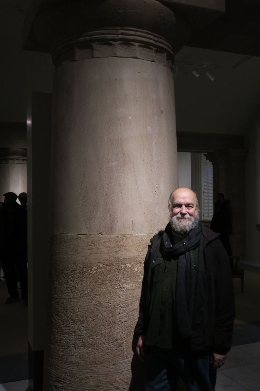 A smiling man with a silver beard, dressed in black, stands in front of a rather large column. The lighting is very gothic.